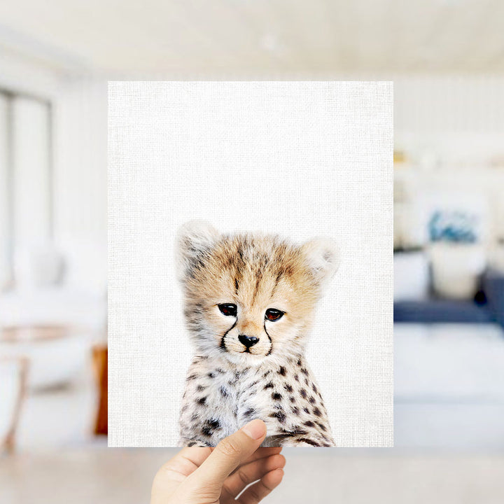A hand is holding up a framed print of a young cheetah with a focused expression, against a blurred background.