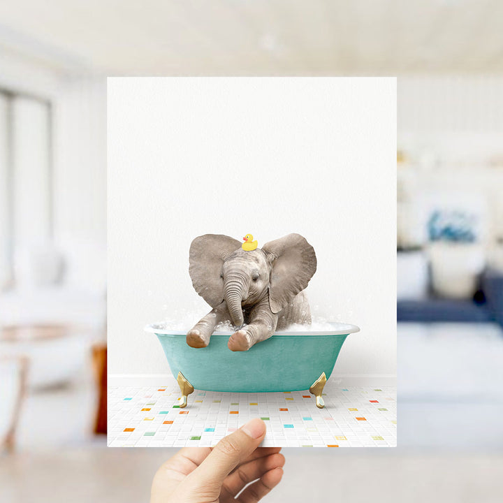 A hand holds up a framed image of a baby elephant sitting in a bathtub filled with bubbles, wearing a yellow rubber duck.