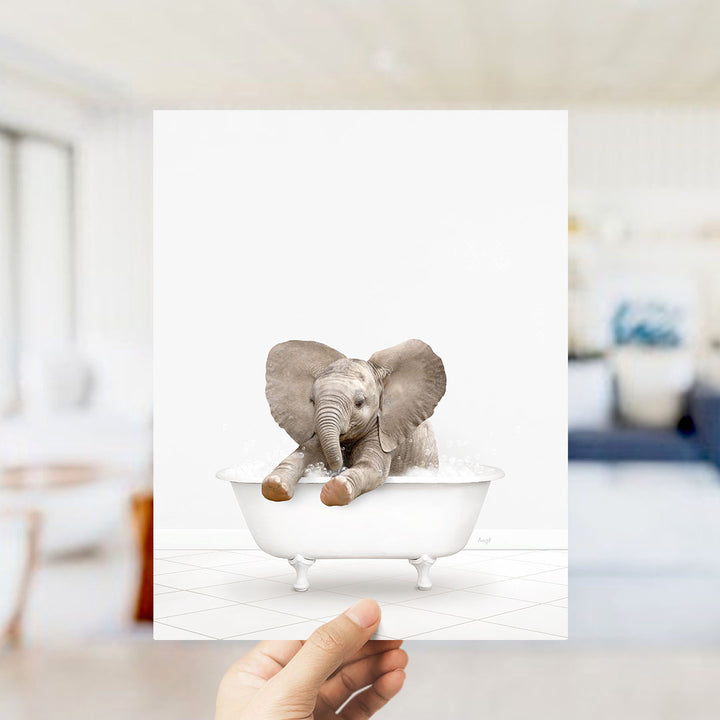 A hand holds up a framed image of a baby elephant sitting in a bathtub, with its trunk raised and ears flapping.