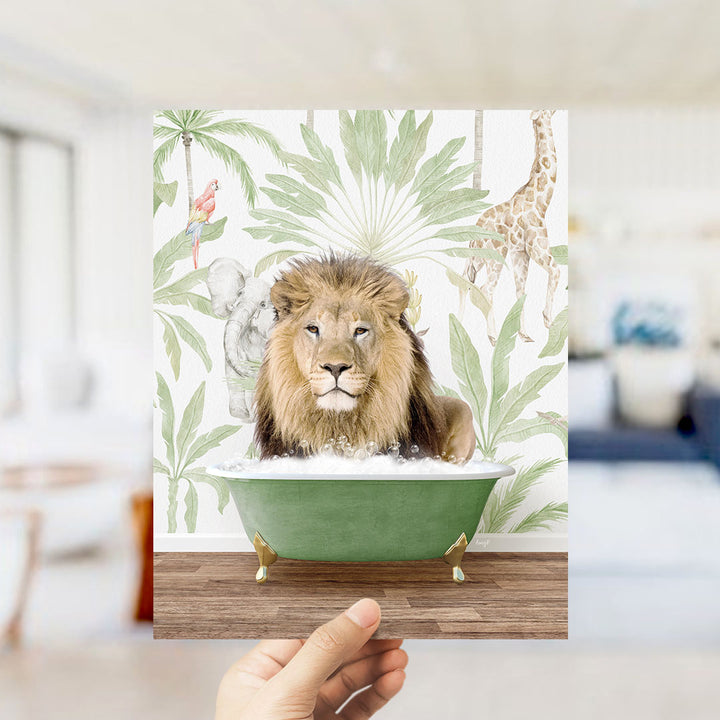 A hand holds up a framed print featuring a lion sitting in a green bathtub surrounded by tropical foliage, including a giraffe and palm trees.