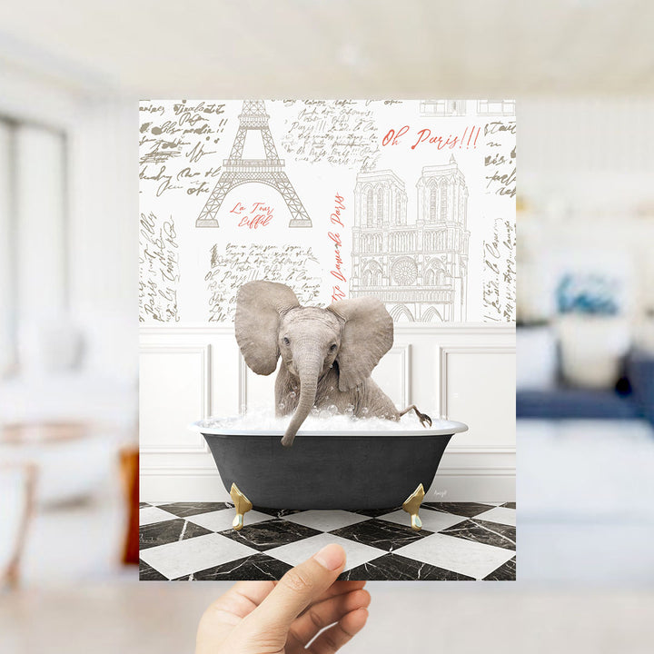 A hand holds up a printed image featuring an elephant sitting in a bathtub, surrounded by various Paris-themed elements such as the Eiffel Tower and the Notre-Dame Cathedral.