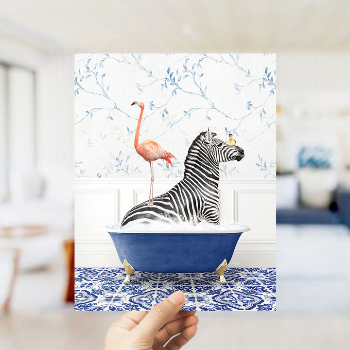 A hand holds up a framed image depicting a zebra sitting in a blue bathtub, with a pink flamingo standing on its back.
