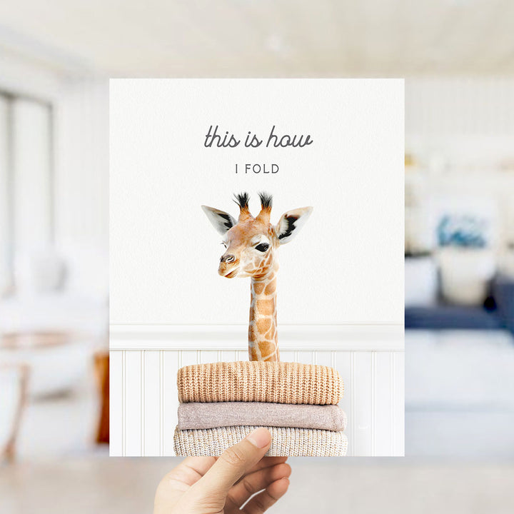 A hand is holding up a white card with a photo of a giraffe standing on a stack of folded towels.