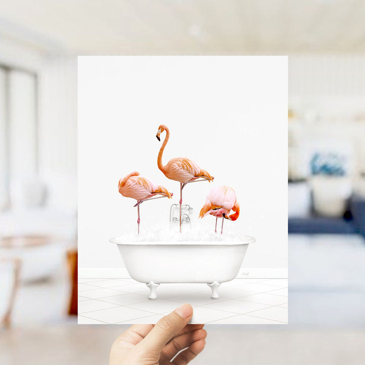A hand holds up a framed image depicting three flamingos standing in a white bathtub filled with water.