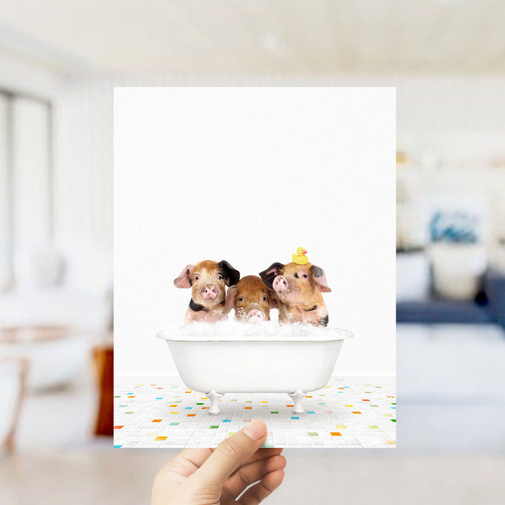 A hand holds up a framed image of three adorable pigs sitting in a bathtub filled with bubbles, with a yellow rubber duck perched on their heads.