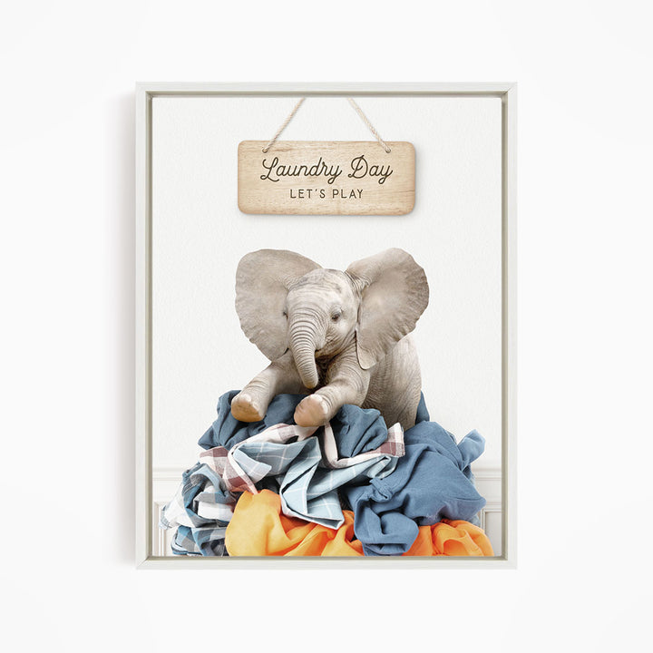 A framed image of a baby elephant sitting atop a pile of laundry, with a sign above that says "Laundry Day Let's Play".