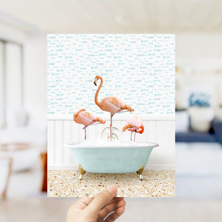 A hand holds up a framed image depicting three flamingos standing in a bathtub filled with bubbles, surrounded by a pattern of fish.