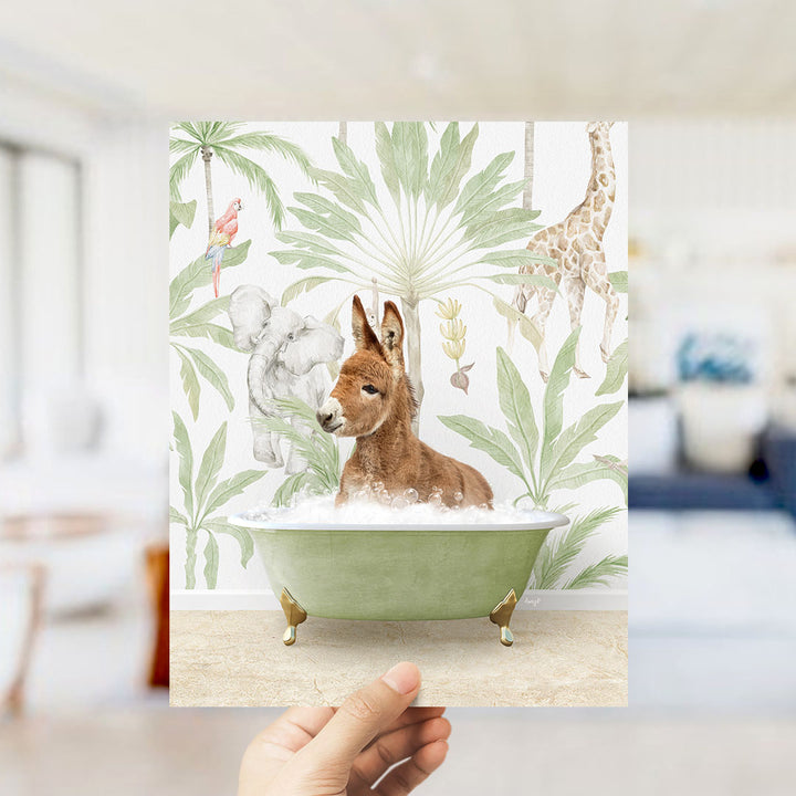 A hand holds up a framed print featuring a whimsical illustration of a donkey sitting in a bathtub surrounded by tropical foliage, including a giraffe and palm trees.