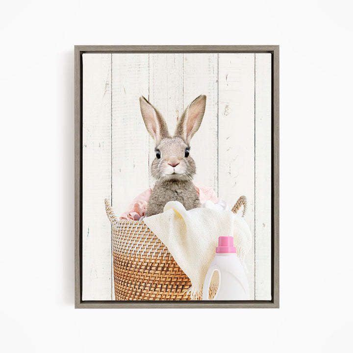 A cute gray rabbit peeking out from a woven basket filled with laundry, including a white towel and a pink bottle.