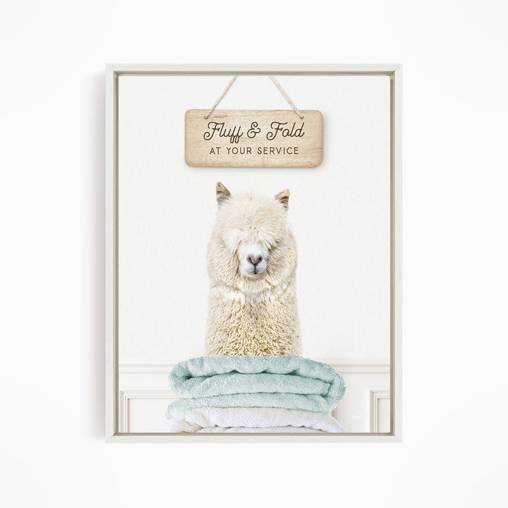 A fluffy white alpaca sitting on a stack of light blue towels, with a wooden sign above that says "Fluffy & Fold at your service".