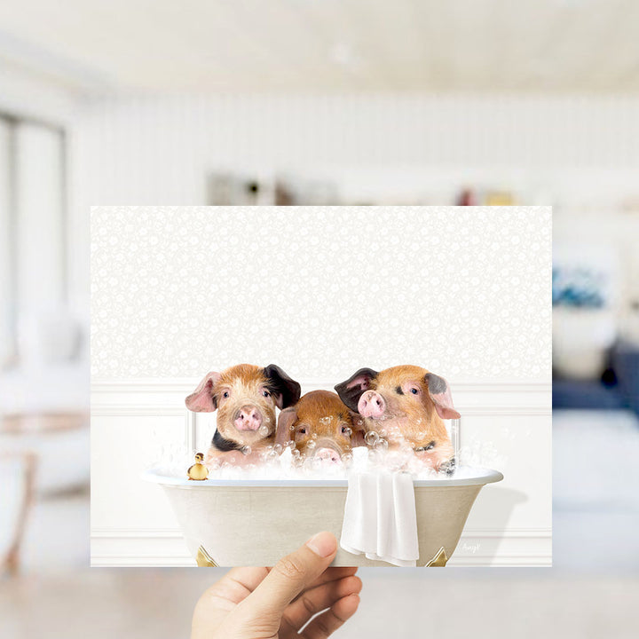 A hand holding a framed image of three adorable pigs sitting in a bathtub filled with bubbles.