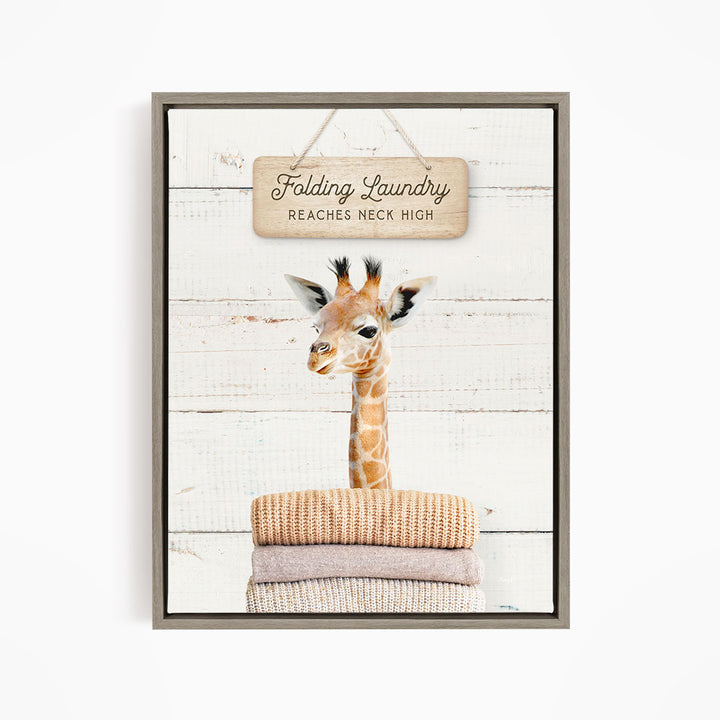 A framed image of a giraffe standing on a stack of folded clothes, with a sign above that says "Folding Laundry Reaches Neck High".