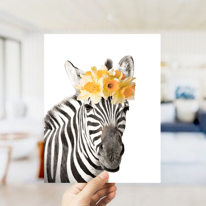 A hand holds up a framed photograph of a zebra with yellow flowers on its head, creating a whimsical and playful scene.