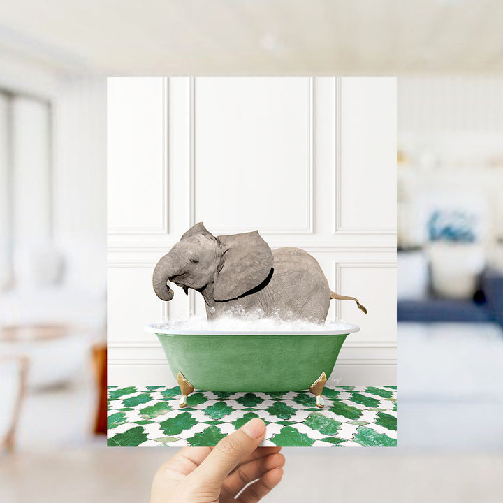 A hand holds up a framed image of a baby elephant sitting in a green bathtub filled with bubbles, surrounded by a green and white patterned floor.