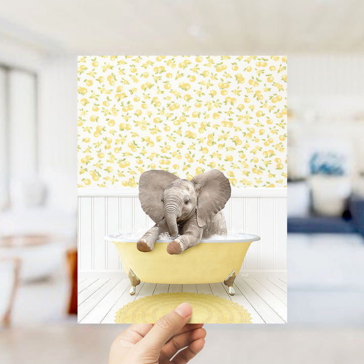 A hand holds up a framed image of a baby elephant sitting in a yellow bathtub, surrounded by a yellow and white floral pattern.