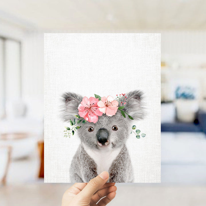 A hand is holding up a framed print of a koala wearing a flower crown.