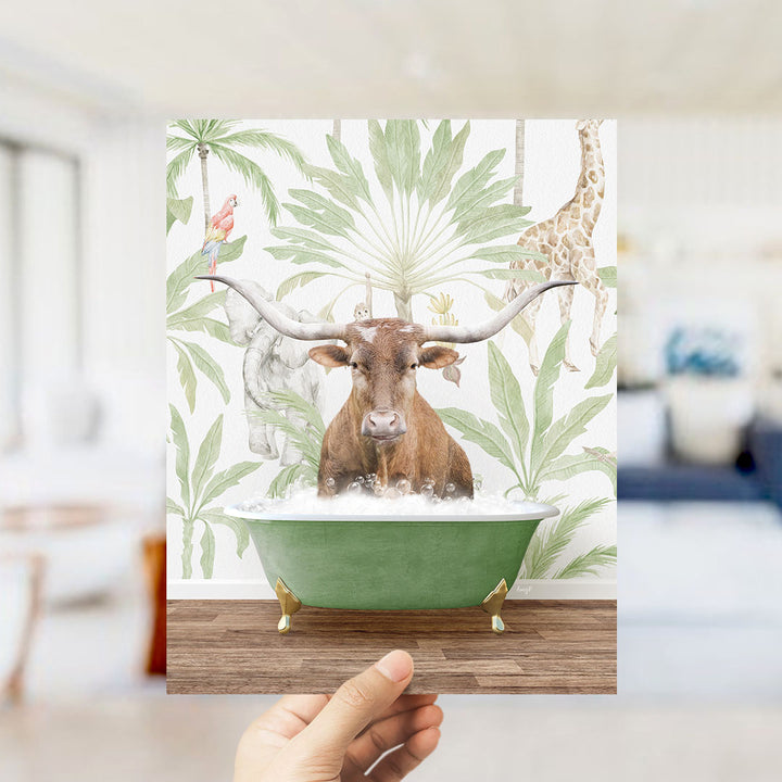 A hand holds up a framed image depicting a brown cow with large horns sitting in a green bathtub, surrounded by tropical foliage and animals.