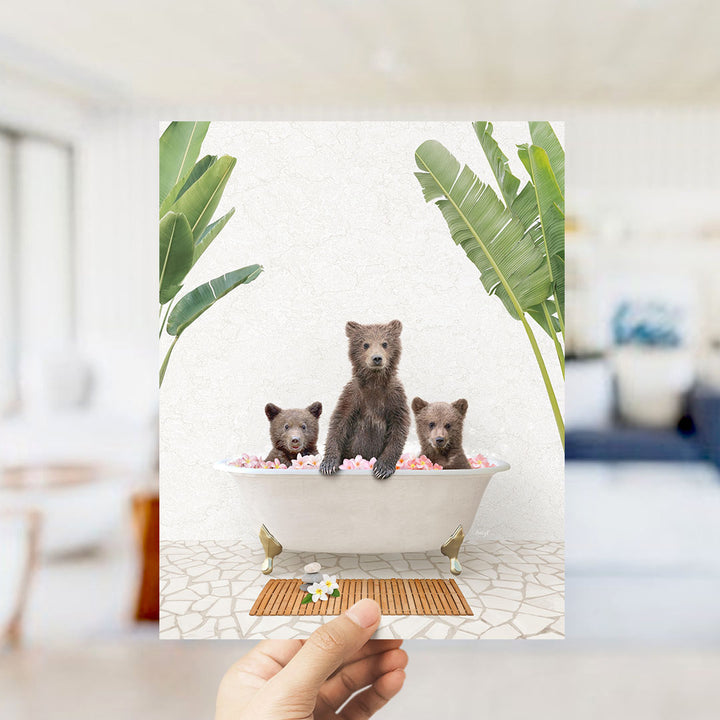 A hand is holding up a framed image depicting three bears sitting in a bathtub filled with pink flowers.