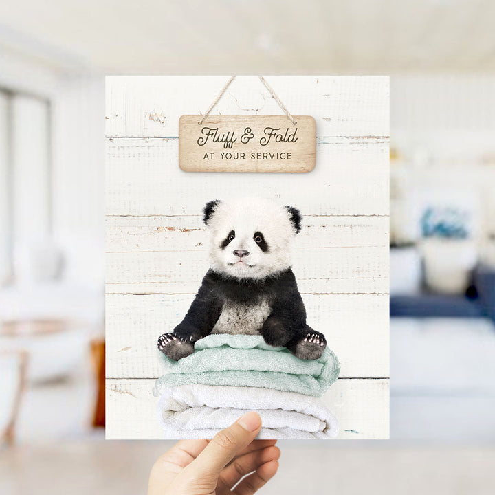 A hand is holding up a card with a cute panda bear sitting on a stack of towels, with a wooden sign above that says "Fluffy & Fold at your service".