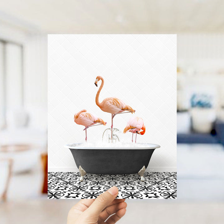 A hand holds up a framed image of three flamingos standing in a bathtub filled with water.