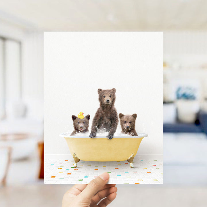 A hand holds up a framed image depicting three adorable brown bears sitting in a yellow bathtub filled with water, surrounded by colorful tiles on the floor.