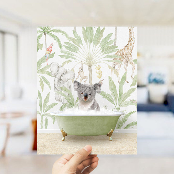 A hand holds up a framed print featuring a koala sitting in a bathtub surrounded by tropical foliage, including a giraffe and other animals.