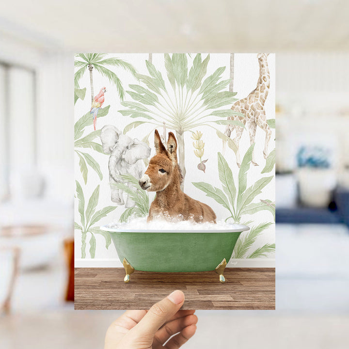 A hand holds up a framed print featuring a whimsical scene of a donkey sitting in a bathtub surrounded by tropical foliage, including a giraffe and palm trees.