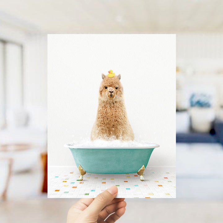 A hand holds up a framed image of a brown alpaca sitting in a blue bathtub filled with bubbles, with a yellow rubber duck perched on its head.