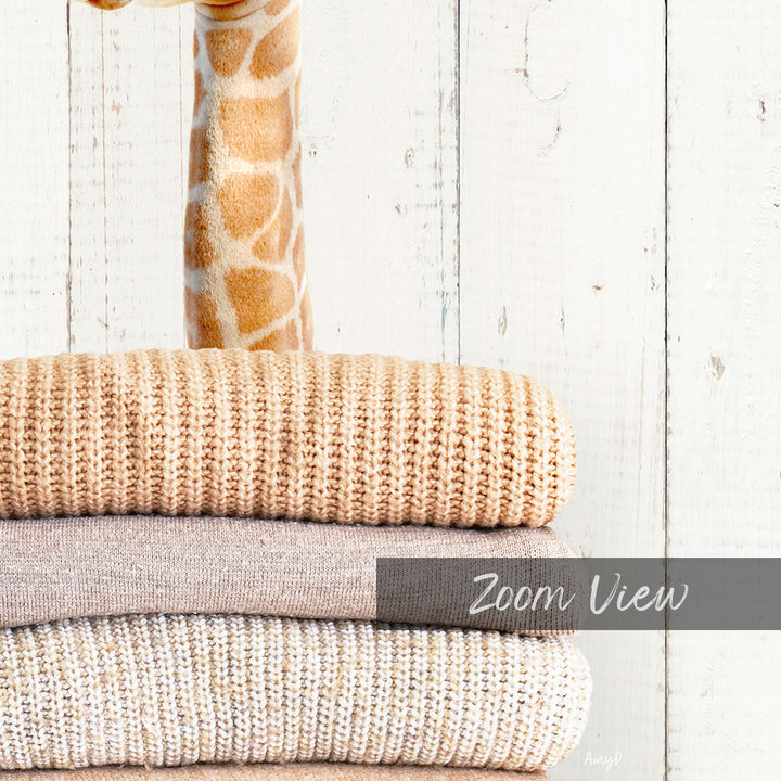 A stack of various beige and gray knitted or woven items, including a blanket and a sweater, against a white wooden background.