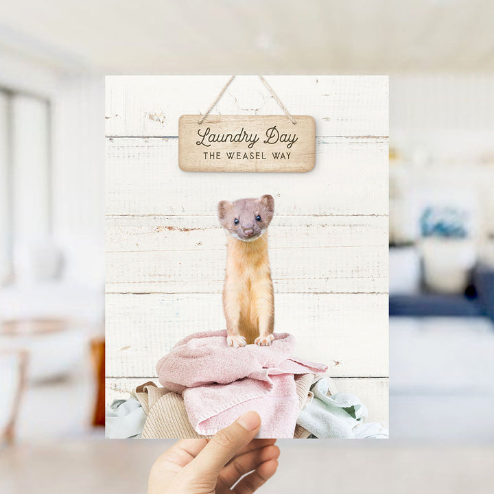 A weasel standing on a pile of clothes, with a wooden sign above it that says "Laundry Day The Weasel Way".