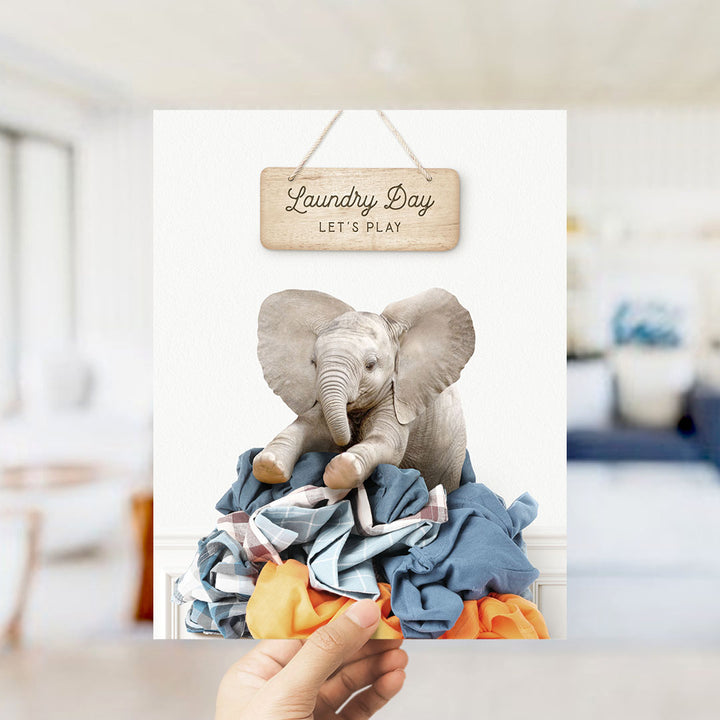 A hand holding a framed image of a baby elephant sitting atop a pile of laundry, with a wooden sign above that says "Laundry Day Let's Play".