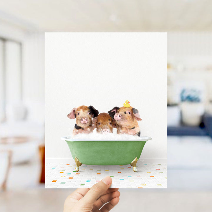 A hand holds up a framed image of three adorable pigs sitting in a green bathtub filled with bubbles, with a yellow rubber duck perched on the side.