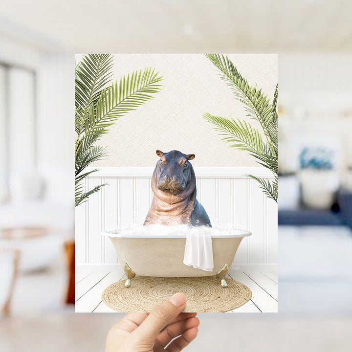 A hand holds up a framed image depicting a hippopotamus sitting in a bathtub filled with water, surrounded by palm leaves.