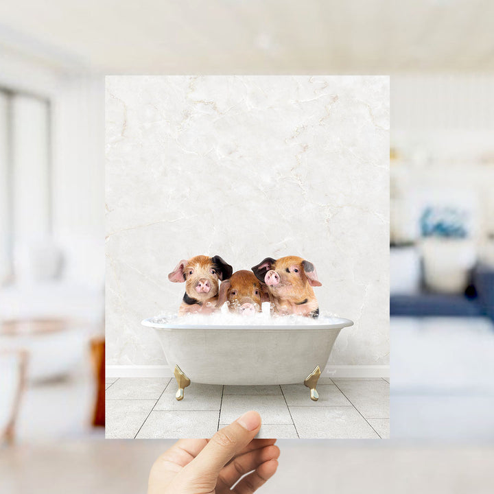 A hand holds up a framed image of three adorable pigs sitting in a bathtub, with a blurred background suggesting a bathroom setting.