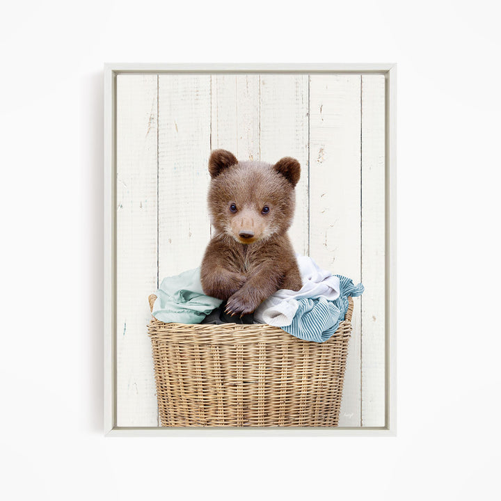 A cute brown bear cub sitting in a wicker basket filled with clothes.