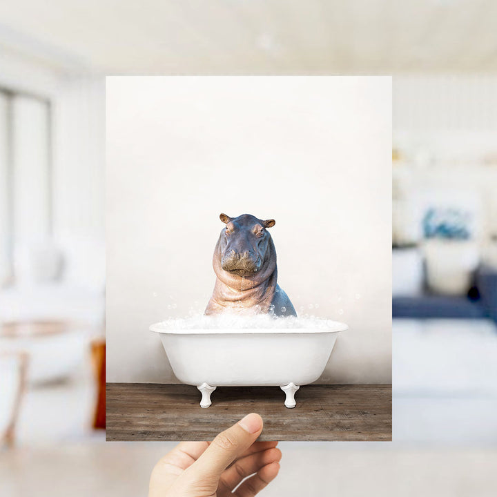 A hand holds up a framed image of a hippopotamus sitting in a bathtub filled with water, creating a whimsical and playful scene.