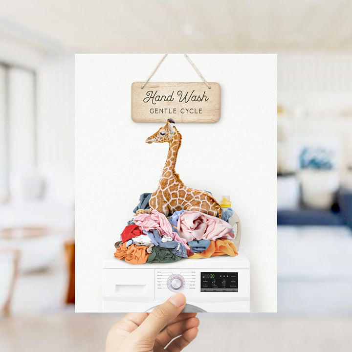 A hand holding a sign that says "Hand Wash Gentle Cycle" above a pile of laundry with a giraffe sitting on top.