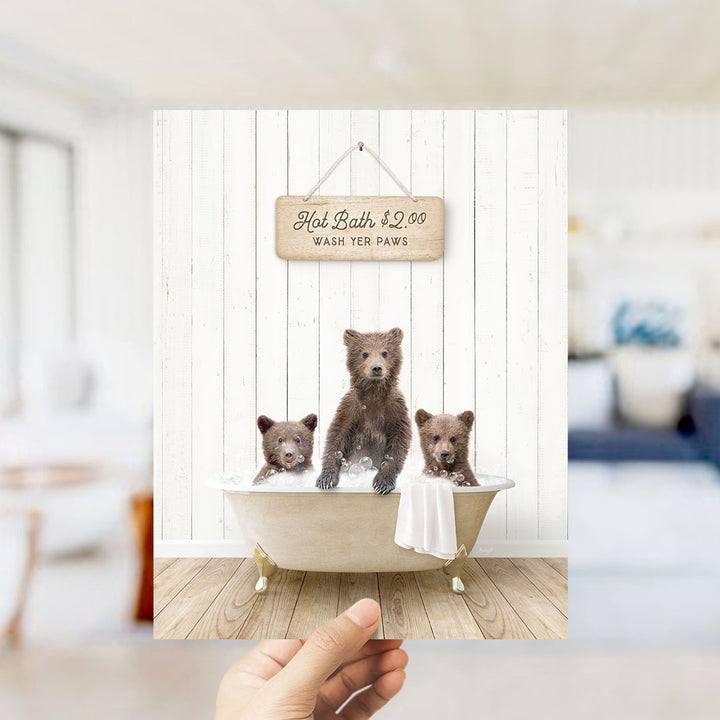 A hand holding a framed image of three bears sitting in a bathtub, with a sign above them advertising hot baths for $2.00 and washing the bear paws.