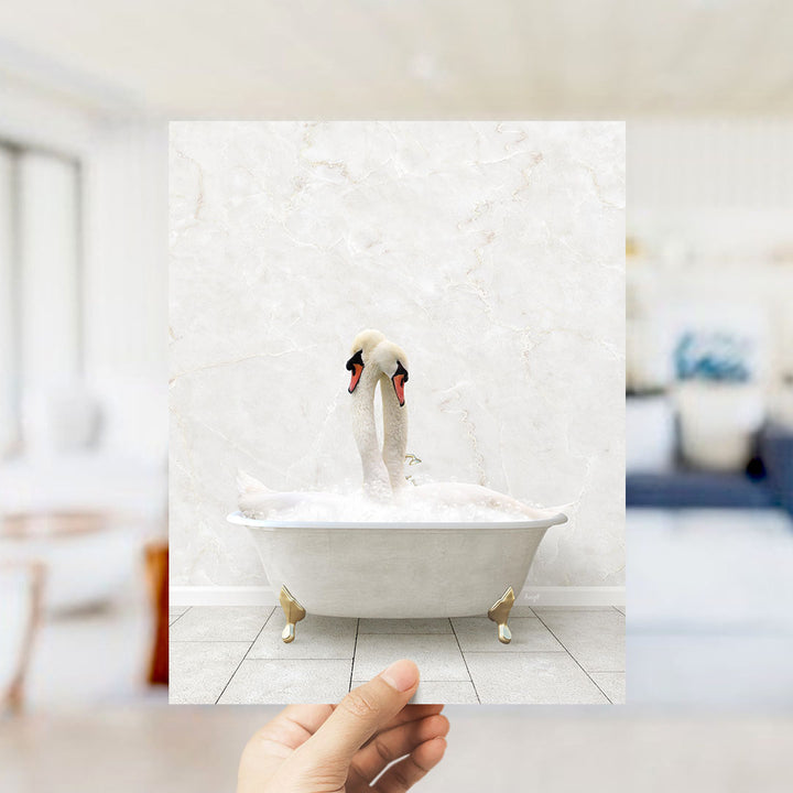 A hand holds up a framed photograph of two swans sitting in a bathtub, with a blurred background suggesting a bathroom setting.