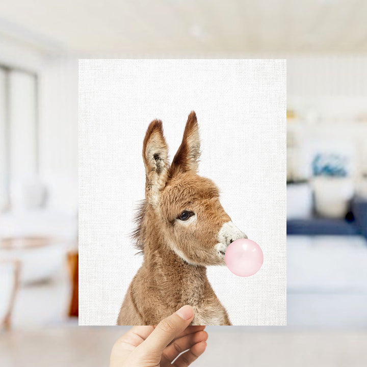 A hand is holding up a picture of a donkey blowing a bubble with a pink bubble gum bubble.