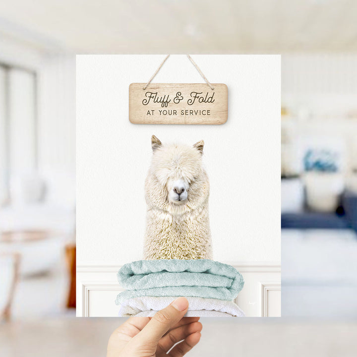 A hand holding a sign that says "Fluffy & Fold at your service" above a photo of a fluffy white alpaca.