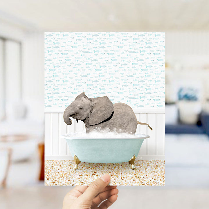 A hand holds up a framed image depicting a baby elephant sitting in a bathtub filled with bubbles, surrounded by a pattern of small fish.