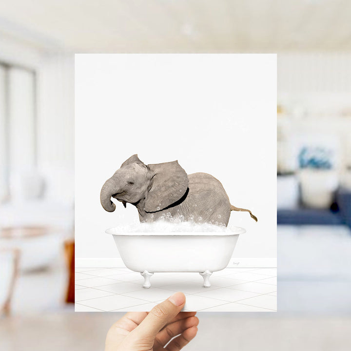 A hand holds up a framed image depicting a baby elephant sitting in a bathtub filled with water.