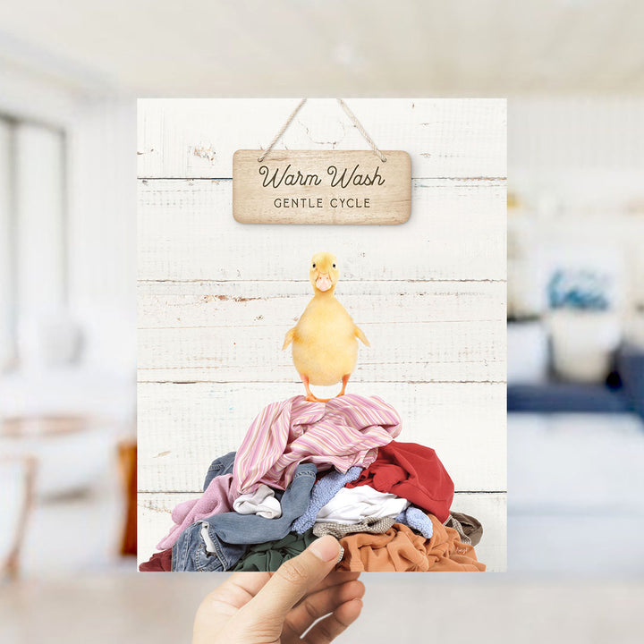 A hand holding a sign with a yellow duck standing on a pile of laundry, with a wooden sign above it that says "Warm Wash Gentle Cycle".