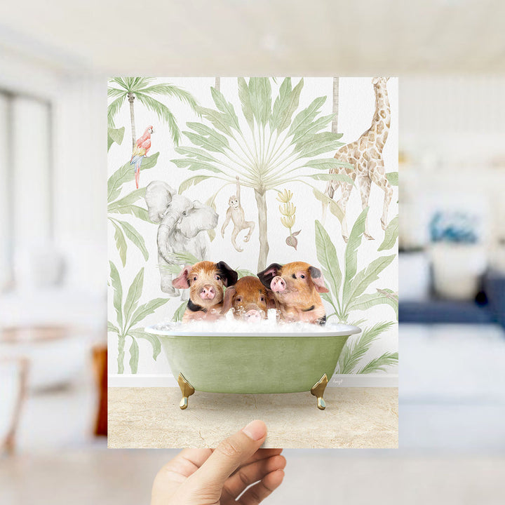 A hand holds up a framed print featuring three pigs sitting in a green bathtub, surrounded by a tropical jungle scene with various animals and plants.