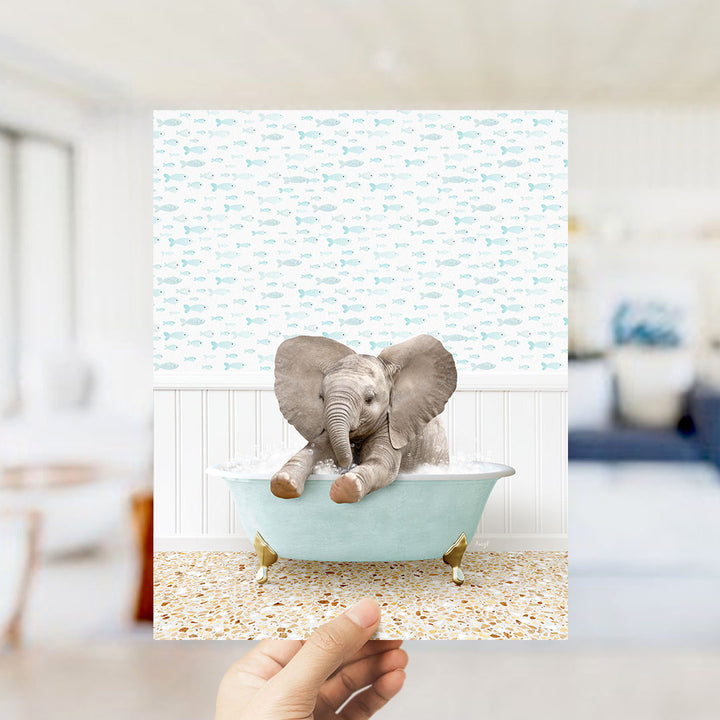 A hand holds up a framed image of a baby elephant sitting in a bathtub filled with bubbles, surrounded by a pattern of small fish.