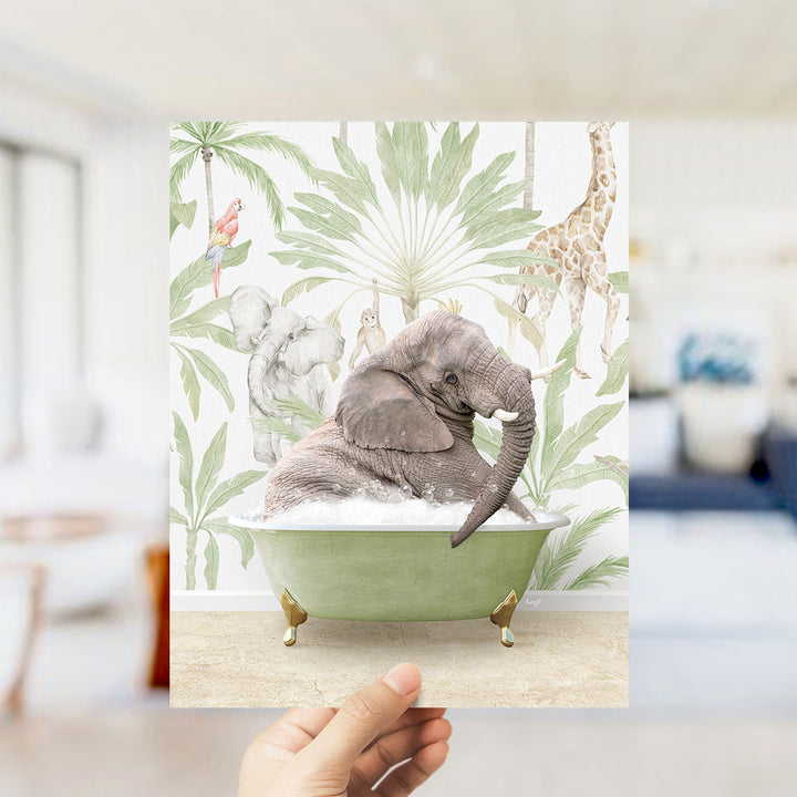 A hand holds up a framed print featuring an elephant sitting in a bathtub surrounded by tropical foliage, including a giraffe and palm trees.