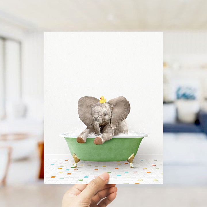 A hand holds up a framed image of a baby elephant sitting in a green bathtub, with a yellow rubber duck on its head.
