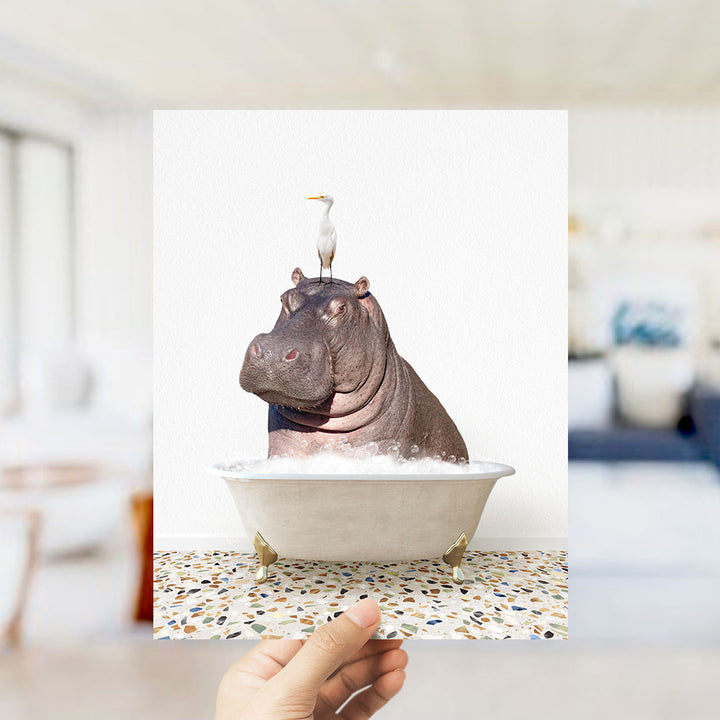 A hand holds up a framed image of a hippopotamus sitting in a bathtub, with a seagull perched on its head.