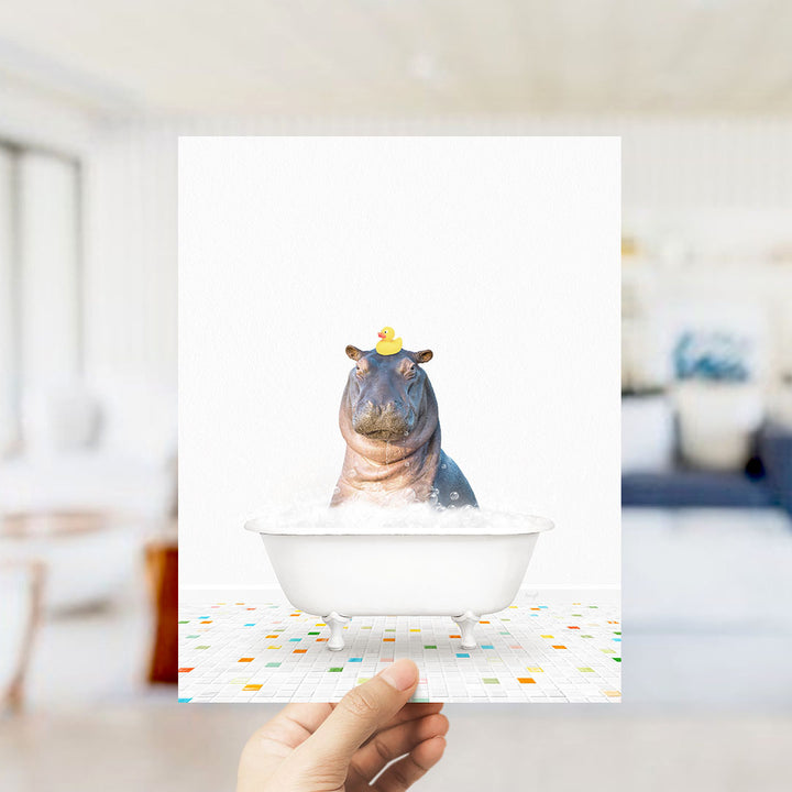 A hand holds up a framed image depicting a hippopotamus sitting in a bathtub, with a yellow rubber duck floating on its head.